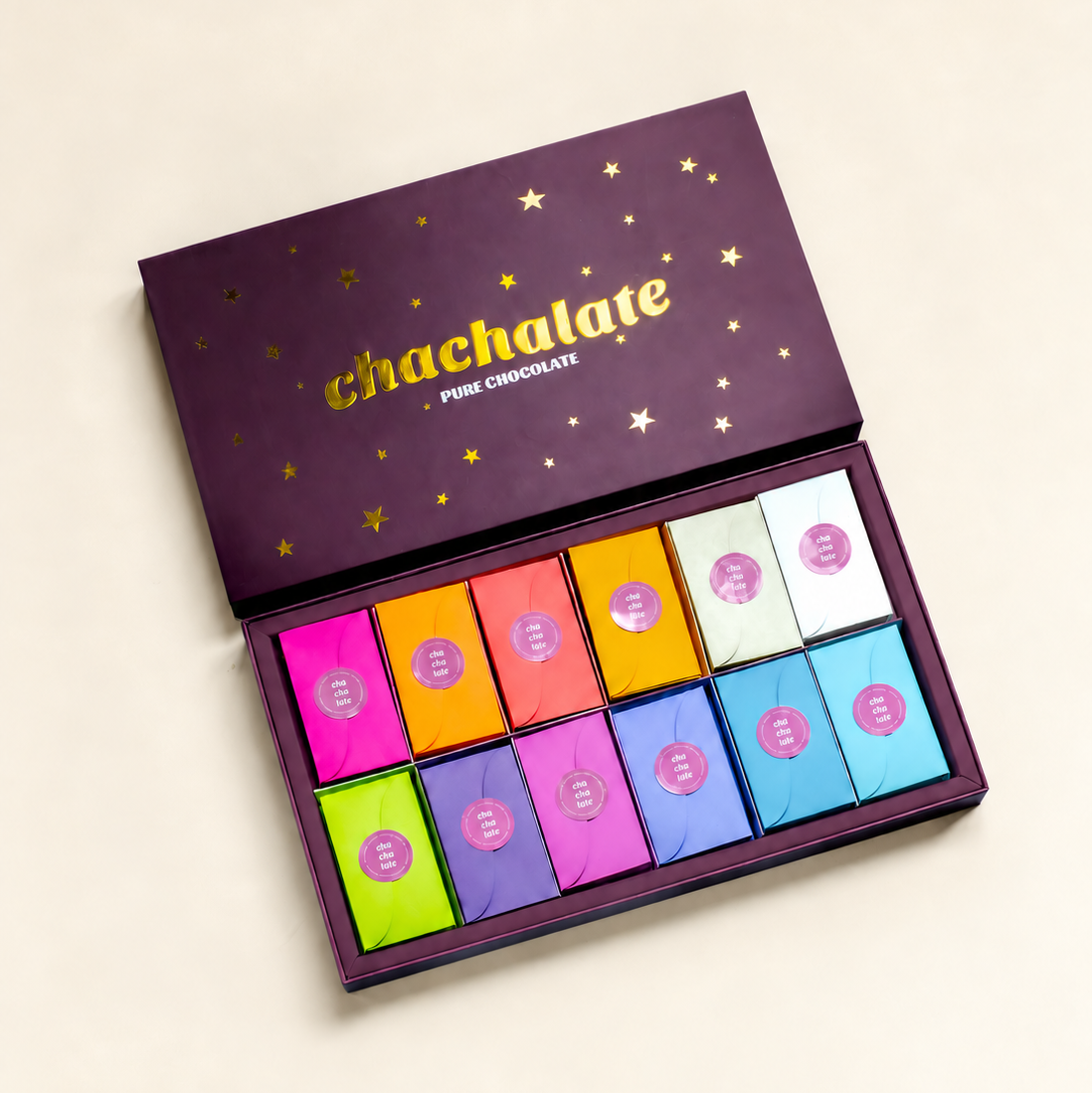 An open purple box of chachalate pink chocolate, showing twelve individually wrapped chocolate squares in a grid of vibrant colors including pink, orange, yellow, green, purple, blue, and white. The box lid features gold star illustrations and the brand name 'chachalate' in gold script.